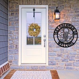 May include: A black metal welcome sign with the text "1583 Sherman Dr Welcome" and a desert scene with a cactus and mountains. The sign is mounted on a stone wall next to a white door with a wreath.