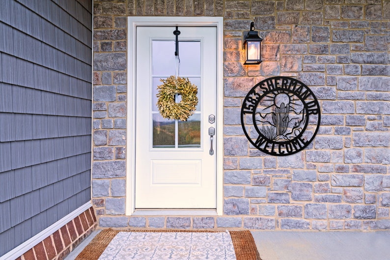 May include: A black metal welcome sign with the text "1583 Sherman Dr Welcome" and a desert scene with a cactus and mountains. The sign is mounted on a stone wall next to a white door with a wreath.