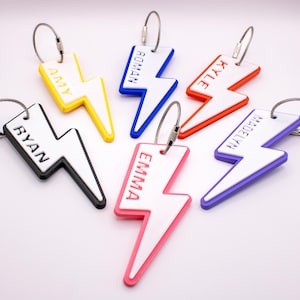 May include: A collection of personalized lightning bolt-shaped keychains in various colors, including yellow, blue, red, pink, purple, and black. Each keychain features a different name in a contrasting color, attached to a silver metal loop.