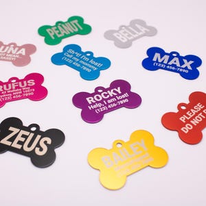 May include: A selection of colourful, bone-shaped pet ID tags. Each tag displays a different name, like "Peanut" or "Max", with some including contact details. The tags are in various colours, including pink, blue, and yellow.