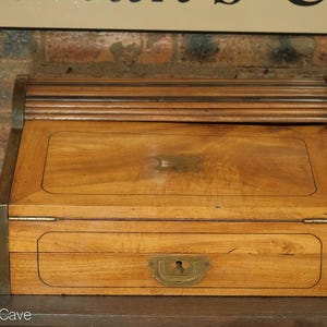 May include: A wooden writing desk with a roll-top lid and a brass lock. The desk has a light brown finish and is decorated with a dark brown inlay.