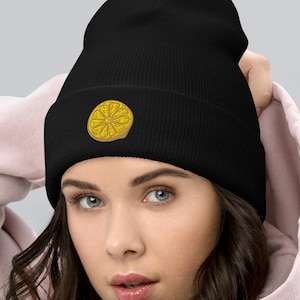 Może przedstawiać: Czarna czapka beanie ze złotym, haftowanym okrągłym wzorem. Czapka wykonana jest z miękkiego materiału i noszona jest na głowie. Ta czapka to stylowy dodatek na chłodne dni i można ją łączyć z różnymi strojami.