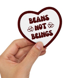 Könnte beinhalten: Ein weißer herzförmiger Patch mit einer roten gestickten Botschaft, die "BEANS NOT BEINGS" lautet, und zwei kleinen Bohnenillustrationen.