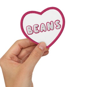 Puede incluir: Un parche en forma de corazón rosa con costuras blancas alrededor de los bordes. La palabra "BEANS" está escrita en letras blancas en el parche.