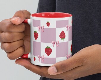 May include: A white ceramic mug with a red rim and handle. The mug features a pink and white checkered pattern with red strawberries and pink bows.