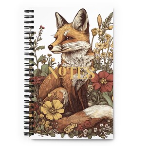 Può includere: Quaderno a spirale con un'illustrazione dettagliata di una volpe in mezzo a fiori colorati. La volpe è marrone e arancione, con la parola "NOTES" in oro. Il quaderno ha una copertina bianca.