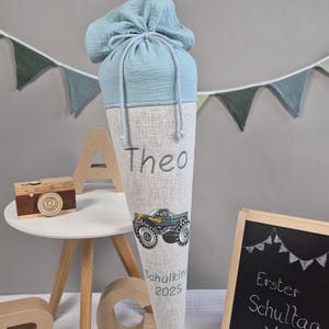 Könnte beinhalten: Eine hellblaue und beige Schultüte mit dem Namen "Theo", einem Monster-Truck-Motiv und dem Text "Schulkin 2025". Die Tüte ist mit hellblauem Stoff und einem Kordelzug verschlossen. Eine kleine Tafel zeigt "Erster Schultag".