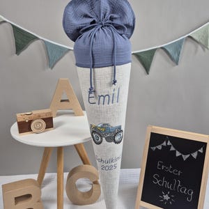 Könnte beinhalten: Eine blau-weiße Schultüte mit dem Namen "Emil" und einem Monster-Truck-Motiv. Die Tüte wird von einem blauen Stoffbeutel gekrönt. Eine kleine Tafel zeigt "Erster Schultag".