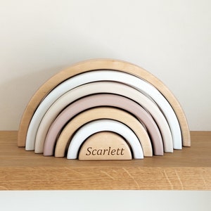 Op de afbeelding: Een houten regenboog stapel speelgoed met vijf bogen in beige, wit en roze tinten. De onderste boog is gegraveerd met de naam "Scarlett".
