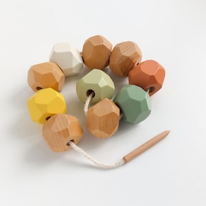 May include: A colorful wooden bead garland with geometric shapes in various colors, including yellow, green, and orange. The beads are strung on a white cord with a wooden peg at the end. The garland is designed for decorative purposes.