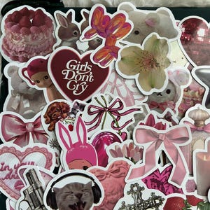 May include: A collection of pink and white stickers featuring various designs, including a bunny, a cat, a flower, a heart, a bow, a cross, and the text "Girls Don't Cry".
