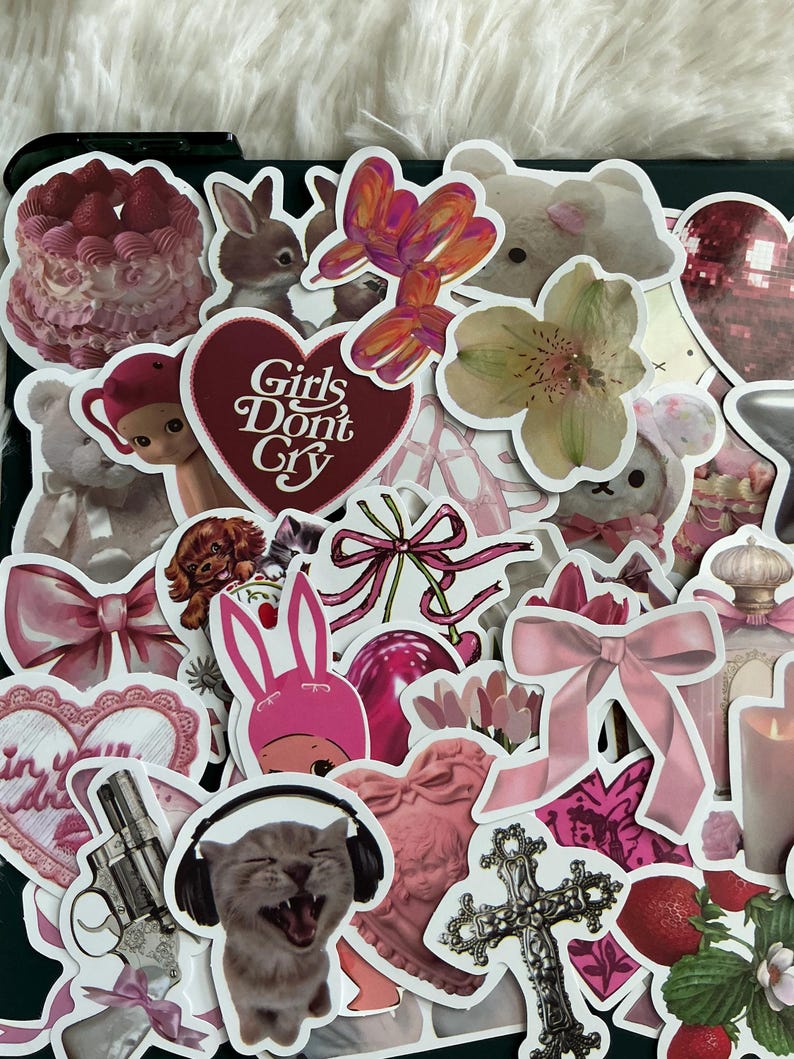 May include: A collection of pink and white stickers featuring various designs, including a bunny, a cat, a flower, a heart, a bow, a cross, and the text "Girls Don't Cry".