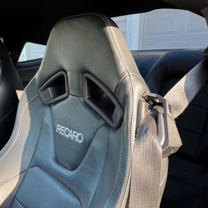 May include: Black leather car seats with the Recaro logo in silver. The seats have integrated headrests and side bolsters for support. A seatbelt is visible on the right side of the image. The interior of a car is visible.