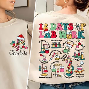 Op de afbeelding: Beige sweatshirt met een kleurrijke afbeelding op de achterkant met de tekst "12 Days of Lab Work" met illustraties van laboratoriumapparatuur. De voorkant heeft een DNA-streng met een kerstmuts en de naam "Charlotte".