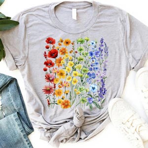Puede incluir: Una camiseta gris claro con un diseño floral vibrante dispuesto en un degradado arcoíris. El diseño presenta una variedad de flores coloridas, incluyendo amapolas rojas, margaritas amarillas y lavanda púrpura. La camiseta está hecha de material suave.