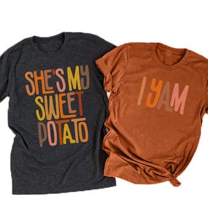 以下が含まれることがあります： 2枚のTシャツ。1枚は濃いグレー、もう1枚は錆色。濃いグレーのシャツには、オレンジ、黄色、茶色で「SHE'S MY SWEET POTATO」と書かれています。錆色のシャツには、オレンジ、茶色、ピンクで「I YAM」と書かれています。