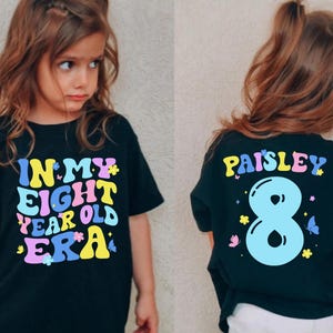 Puede incluir: Camiseta negra con texto colorido que dice "In My Eight Year Old Era" y un número ocho azul con mariposas. La parte posterior de la camiseta dice "Paisley" en letras de colores.