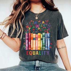 May include: A dark grey t-shirt featuring a graphic of a stack of books with words such as "justice", "love", "compassion", "inclusion", "peace", "unity", "change", "kindness", "diversity", and "hope" printed on the spines. The books are surrounded by colourful flowers and butterflies. The words "EQUALITY" are printed below the books in rainbow colours.