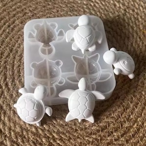 Pode incluir: Um molde de silicone translúcido com quatro cavidades em forma de tartarugas marinhas, acompanhado por três figuras brancas e sólidas em forma de tartaruga moldadas, exibindo padrões de carapaça detalhados e superfícies lisas.