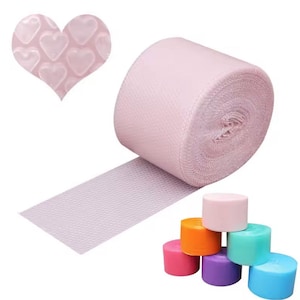 Puede incluir: Un rollo de cinta de tela texturizada de color rosa claro con una superposición de plástico de burbujas en forma de corazón en la esquina superior izquierda y un pequeño grupo de seis rollos de cinta adicionales en rosa, naranja, morado, turquesa y azul.