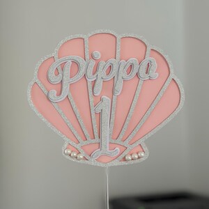 Op de afbeelding: Een roze en zilveren schelpvormige taarttopper met de naam "Pippa" en het cijfer "1". De topper heeft een glinsterende zilveren omtrek en is versierd met kleine parels aan de basis. Het wordt vastgehouden door een dunne zilveren draad.