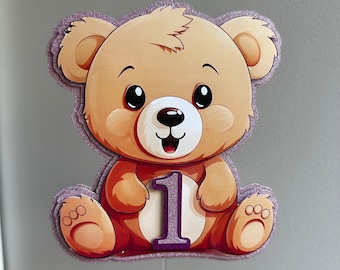 May include: A cartoon-style teddy bear cake topper. The bear is light brown with a purple glitter outline and the number "1" in purple. The bear has large black eyes and is smiling. The topper is attached to a clear plastic stick.