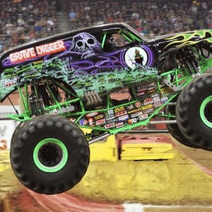 May include: A black and green monster truck, the "Grave Digger," is airborne, displaying a skull graphic and the name on the side. The truck has large black tires with green rims and a green chassis. A black flag with a skull flies from the side.
