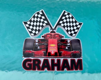 May include: A red Formula 1 race car cake topper with the name "GRAHAM" in bold letters. The topper features two checkered flags and is set against a teal background.