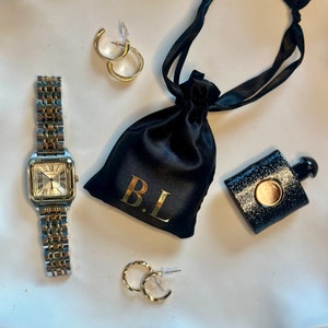 Op de afbeelding: Een zilveren horloge met een witte wijzerplaat en Romeinse cijfers voor de uren, een zwarte satijnen tas met de initialen "B.L." geborduurd in goud, en twee paar gouden oorringen.