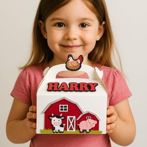 Può includere: Una scatola bianca per bomboniere con un design di fienile rosso, una mucca e un maiale. Il nome "HARRY" è visualizzato in lettere rosse. Un pollo è in cima alla maniglia. La scatola è tenuta da una persona.
