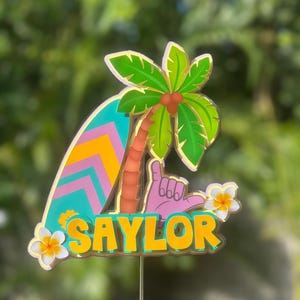 Könnte beinhalten: Ein farbenfroher Tortenaufsatz mit einem Surfbrett, einer Palme und dem Namen "SAYLOR" in Gelb. Das Design beinhaltet eine Hand, die das Shaka-Zeichen macht, sowie weiße und gelbe Blumen. Der Aufsatz befindet sich auf einem dünnen Metallstab.