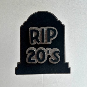 May include: Black tombstone-shaped sign with the words "RIP 20's" in white lettering. The sign is on a stick.