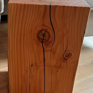 Puede incluir: Un bloque de madera rectangular macizo, probablemente una pequeña mesa o taburete. La madera tiene un tono marrón rojizo cálido con patrones de vetas y nudos prominentes. Grietas oscuras recorren la superficie, añadiendo carácter. La pieza es robusta y parece hecha a mano.