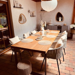 Könnte beinhalten: Ein Esstisch aus Holz mit einem dunklen Metallrahmen, umgeben von geflochtenen Stühlen mit cremefarbenen Kissen. Der Tisch ist mit Tischsets und Glaswaren gedeckt. Ein kleiner geflochtener Hocker steht daneben. Dekorative Papierlaternen hängen von der Decke.