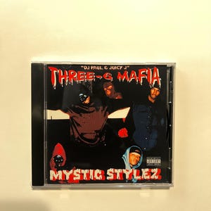 May include: A CD case featuring the album cover for "Mystic Stylez" by Three 6 Mafia. The cover art has a black background with red and white text. The album title is prominently displayed, along with images of the artists.