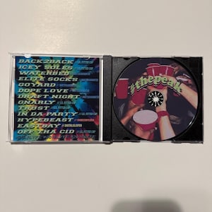 May include: An open CD case displaying a disc with the text "#thepeak" and a list of song titles and artist names. The disc features a graphic of hands holding red cups. The case is on a white surface.
