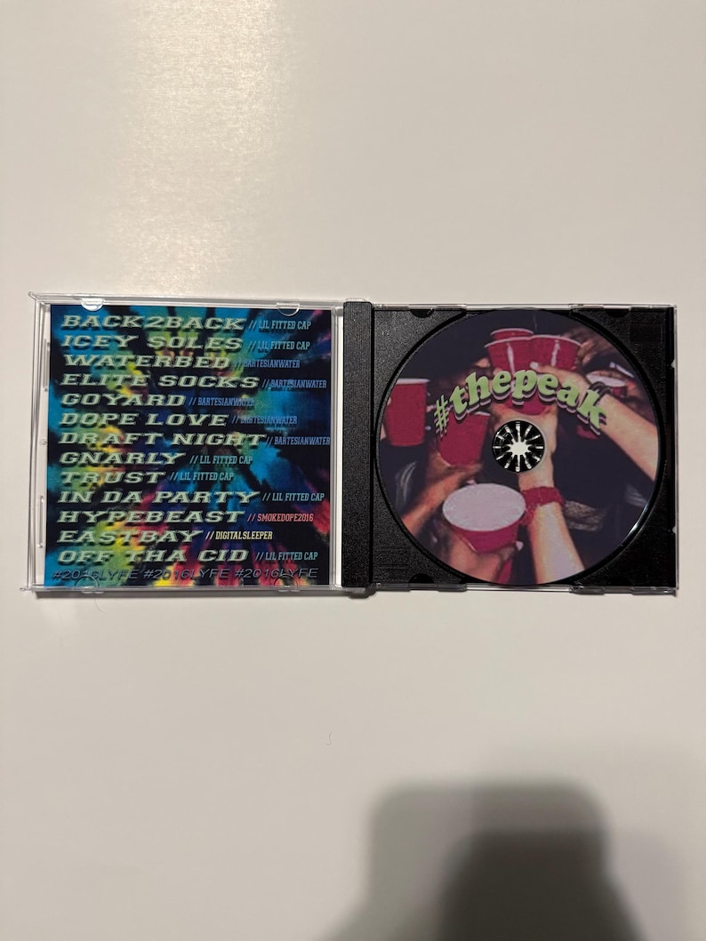 May include: An open CD case displaying a disc with the text "#thepeak" and a list of song titles and artist names. The disc features a graphic of hands holding red cups. The case is on a white surface.