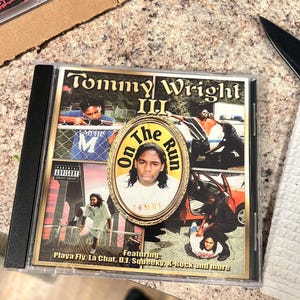 May include: A CD case for Tommy Wright III's album, "On The Run." The cover features a central oval portrait of the artist, surrounded by action shots and the text "Parental Advisory Explicit Content." The CD case is in a clear plastic jewel case.