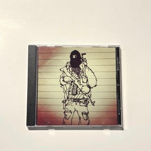 May include: A CD case featuring a black and white illustration of a person in military attire, holding a rifle. The background has horizontal stripes with a gradient of red to yellow. The CD case has a black spine.