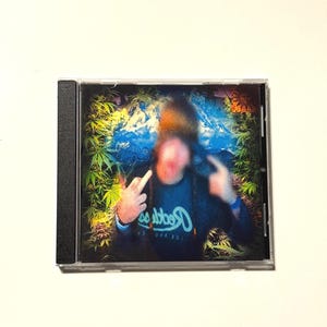 May include: A CD case featuring a blurred image of a person making hand gestures. The background has a blue and green design with cannabis leaves. The person is wearing a dark blue shirt with white text.