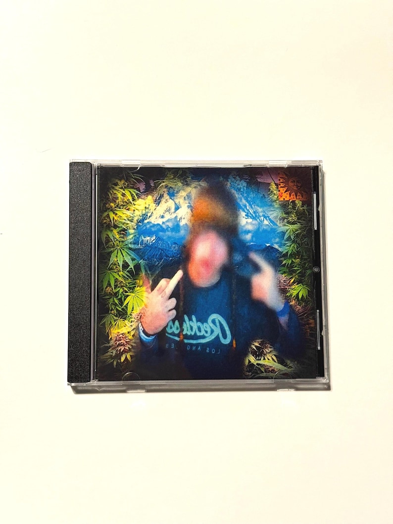 May include: A CD case featuring a blurred image of a person making hand gestures. The background has a blue and green design with cannabis leaves. The person is wearing a dark blue shirt with white text.