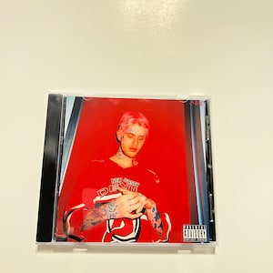 May include: A CD case featuring a portrait of a person with pink hair, wearing a red jersey with "NEW JERSEY DEVILS" printed on it. The image has a red background and a "Parental Advisory Explicit Lyrics" label.
