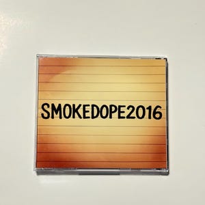 May include: A CD case with a gradient background of orange and yellow horizontal lines. The text "SMOKEDOPE2016" is printed in black across the center of the case.