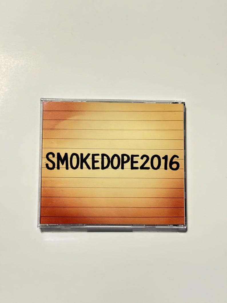 May include: A CD case with a gradient background of orange and yellow horizontal lines. The text "SMOKEDOPE2016" is printed in black across the center of the case.