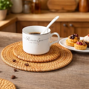 May include: A white ceramic mug with the text "Recherche Snow" and a white spoon sits on a woven rattan coaster. Coffee beans and small pastries are also visible on a wooden surface.