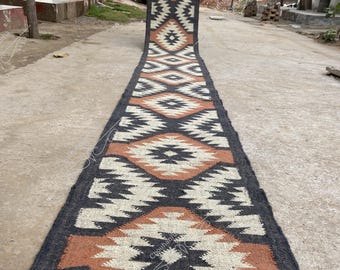 May include: A long, rectangular rug with a geometric pattern. The rug features a repeating diamond design in black, cream, and rust-orange. The rug is made of a woven material and is displayed outdoors on a concrete surface.