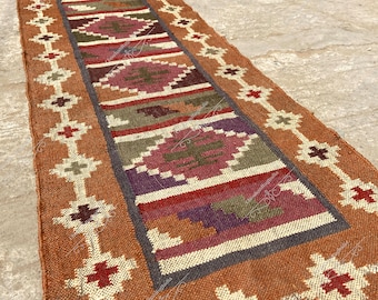 May include: A brown and beige jute rug with a geometric pattern in shades of red, purple, and green. The rug has a fringed edge and is woven with a traditional design.