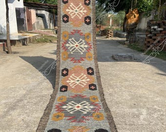 May include: A long, narrow rug with a geometric pattern in shades of gray, red, black, white, and yellow. The rug is woven with a traditional design and has a fringed edge.