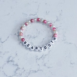May include: A white beaded bracelet with pink and white beads. The beads spell out the word "BEARTOOTH".