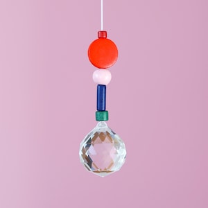 Könnte beinhalten: Ein hängendes Kristallprisma mit einer Schnur, das eine Reihe farbiger Perlen aufweist. Die Perlen umfassen eine rote Scheibe, eine rosa Kugel, einen blauen Zylinder und ein grünes Quadrat, über dem facettierten Kristallkugel. Der Hintergrund ist einfarbig rosa.